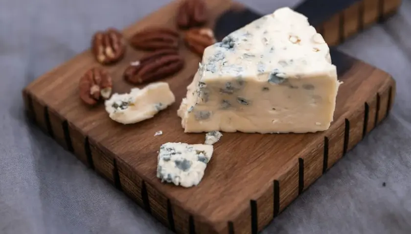Un trozo de queso azul, ideal para la flora intestinal, reposa sobre una tabla de madera junto a nueces pecanas.