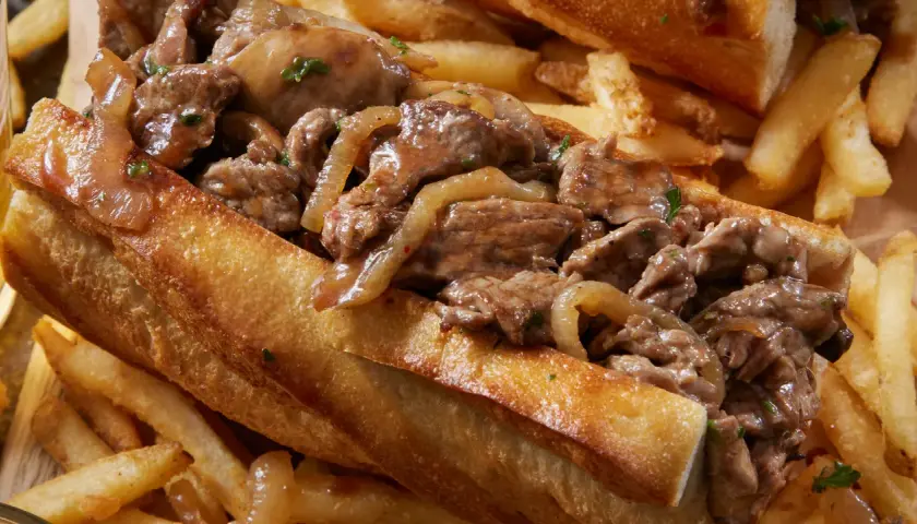 Un delicioso pepito de ternera con cebolla caramelizada, servido en pan crujiente y acompañado de patatas fritas doradas.