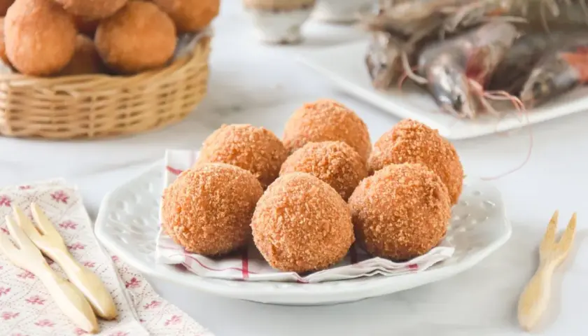 Croquetas de gambas doradas y crujientes en un plato blanco, con más en una cesta de mimbre al fondo.