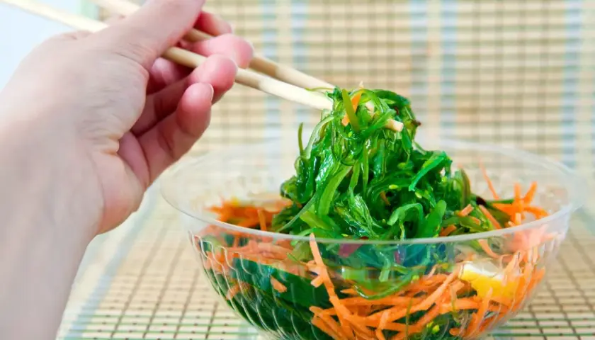 Mano con palillos recogiendo una porción de ensalada de wakame fresca y vibrante, con zanahoria rallada en un bol transparente.
