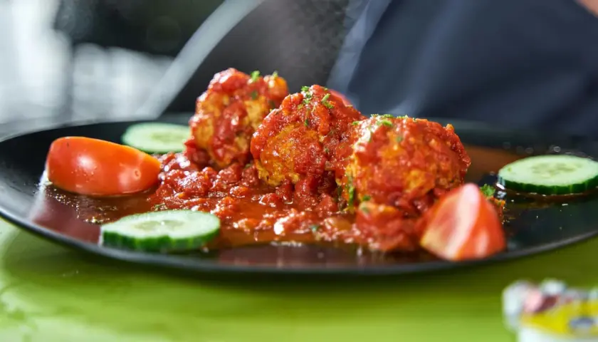 Albondigas con tomate, jugosas y cubiertas de salsa roja, acompañadas de rodajas de pepino y tomate fresco en un plato negro.
