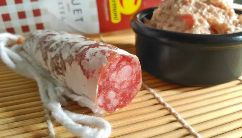 Un trozo de fuet extra con su interior rosado y veteado, junto a un cuenco de paté.