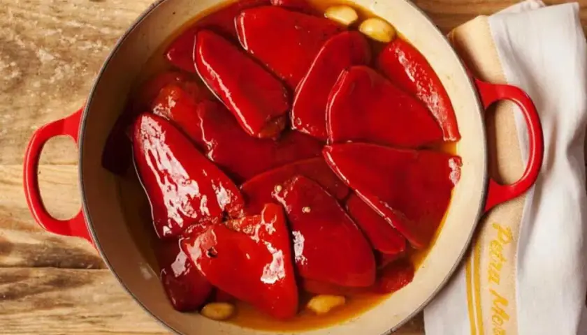 Pimientos del piquillo asados en una cazuela roja, con ajos y un paño de cocina.
