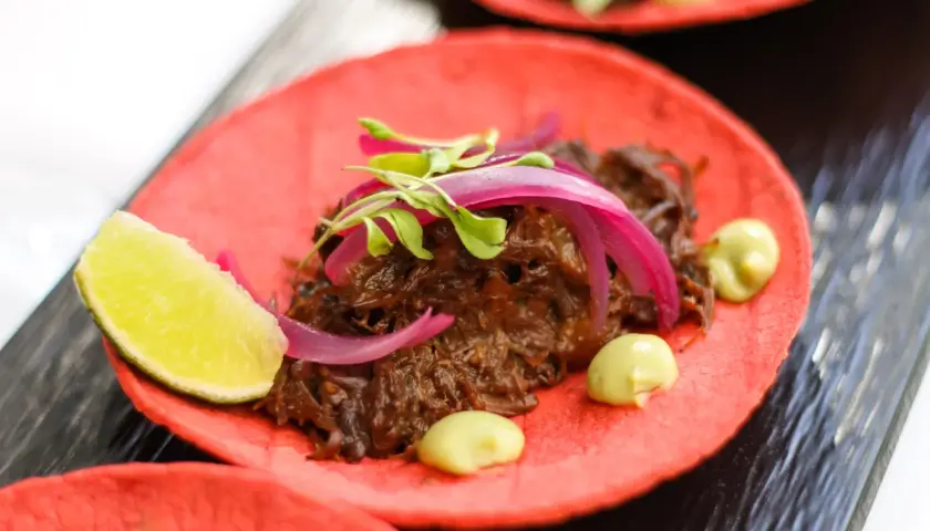 Deliciosos tacos de rabo de toro sobre tortilla roja, con cebolla morada, brotes verdes y un toque de aguacate.