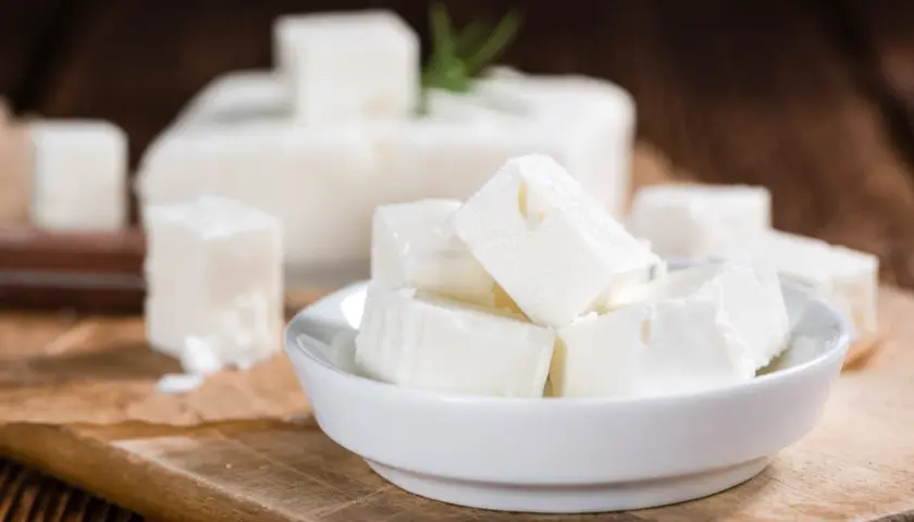 Cubos de queso blanco en un cuenco, con un bloque de queso de leche cruda al fondo. El queso de leche cruda puede presentar peligros.