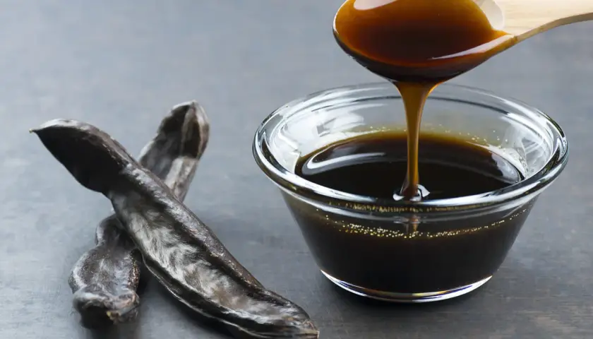 Miel de caña oscura y espesa cayendo de una cuchara de madera a un cuenco de cristal, junto a vainas de algarroba. Descubre sus propiedades.