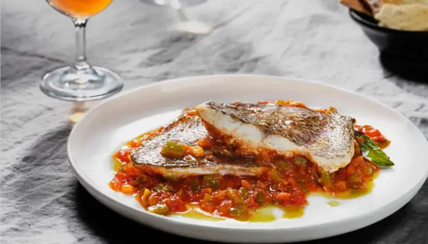 Deliciosa urta a la roteña servida en plato blanco, con guarnición de pimientos y tomate. Acompañada de copa de vino y pan.