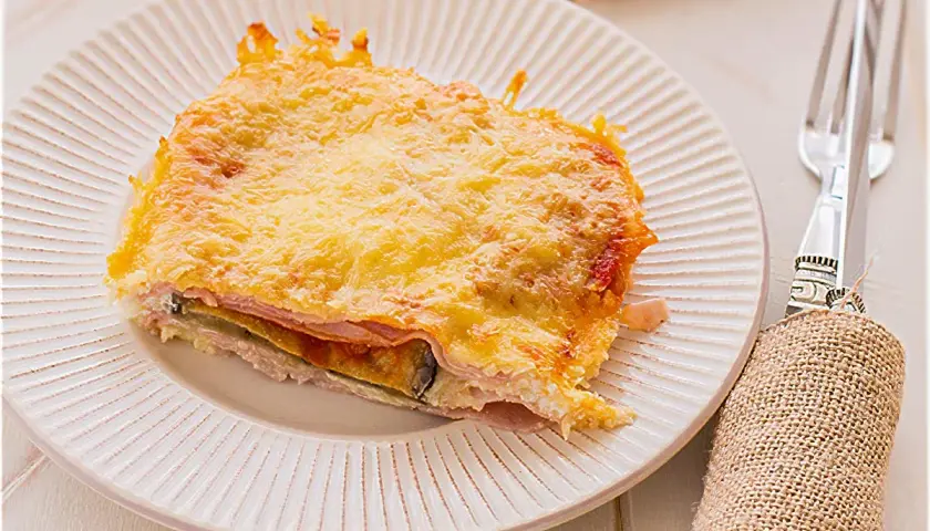 Porción de pastel de berenjenas gratinado con queso, capas de jamón y berenjena, servido en plato blanco.