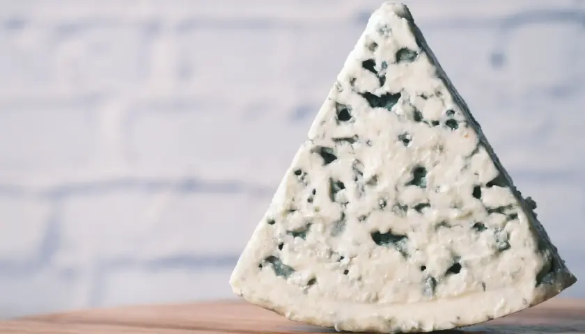 Un trozo triangular de queso azul, con sus características vetas de moho azul verdoso, descansa sobre una superficie de madera.