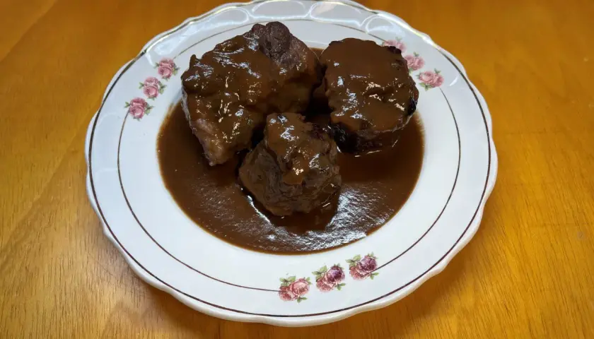 Tres trozos de rabo de toro estofado en una salsa oscura y rica, servidos en un plato blanco con borde dorado y decorado con rosas.