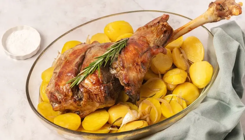 Pierna de cordero al horno dorada, servida con patatas asadas, romero y ajo en una fuente de cristal.