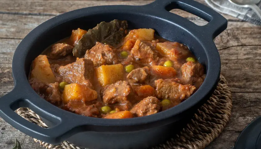 Un reconfortante estofado de ternera con patatas, guisantes y laurel en una cazuela negra sobre madera rústica.