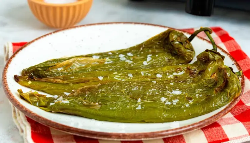 Dos pimientos verdes fritos, salpicados de sal gruesa, reposan en un plato blanco con borde marrón. Un paño a cuadros rojos y blancos y un cuenco de sal completan la escena.