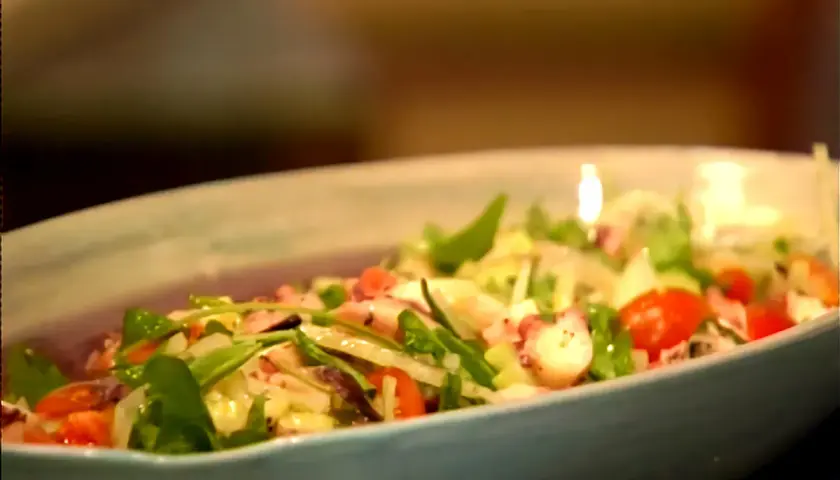 Deliciosa insalata di mare con tentáculos de pulpo, tomates cherry y hojas verdes frescas en un plato azul claro.