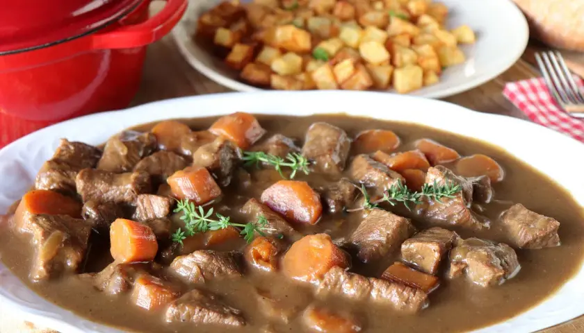 Ternera estofada en salsa con zanahorias y patatas fritas al fondo.
