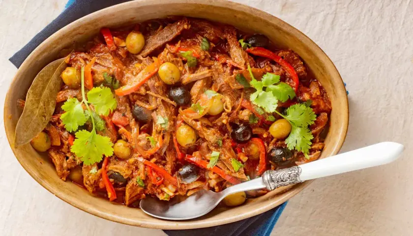 Delicioso plato de ropa vieja con aceitunas verdes y negras, pimientos rojos y cilantro fresco, servido en una cazuela de barro.