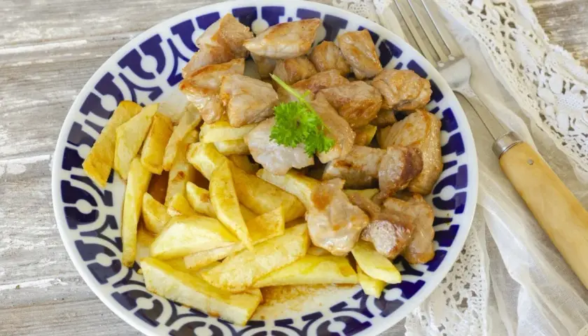 Raxo con patatas, un plato tradicional gallego, servido en un plato azul y blanco con perejil fresco.