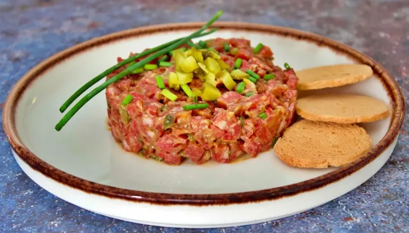 Tartar de salchichón finamente picado, adornado con pepinillos y cebollino, servido con tostas.