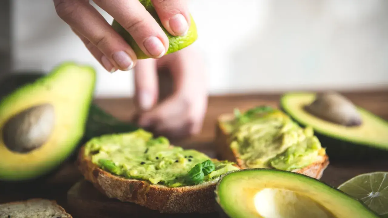 Una mano exprime lima sobre tostadas con aguacate machacado. El aguacate tiene fibra, ideal para un desayuno saludable.
