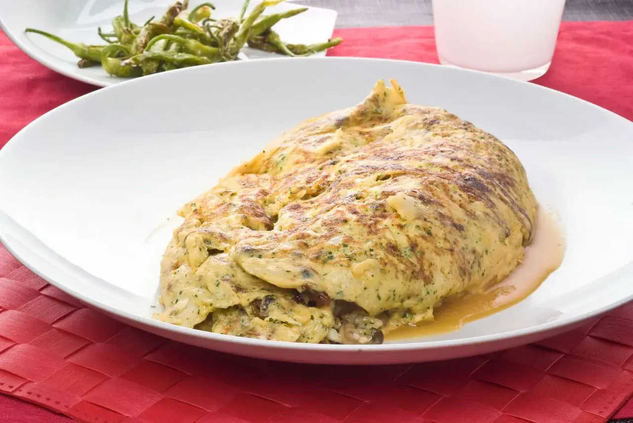 Tortilla de bacalao vasca, dorada y jugosa, servida en plato blanco sobre mantel rojo. Acompañada de pimientos verdes fritos.