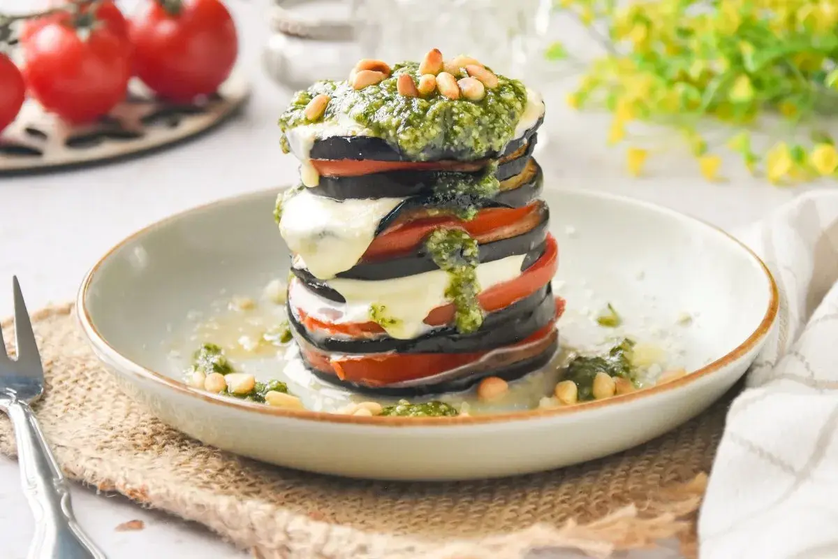 Deliciosa milhoja de berenjena con tomate, mozzarella fundida y pesto, coronada con piñones. Un plato ligero y sabroso.