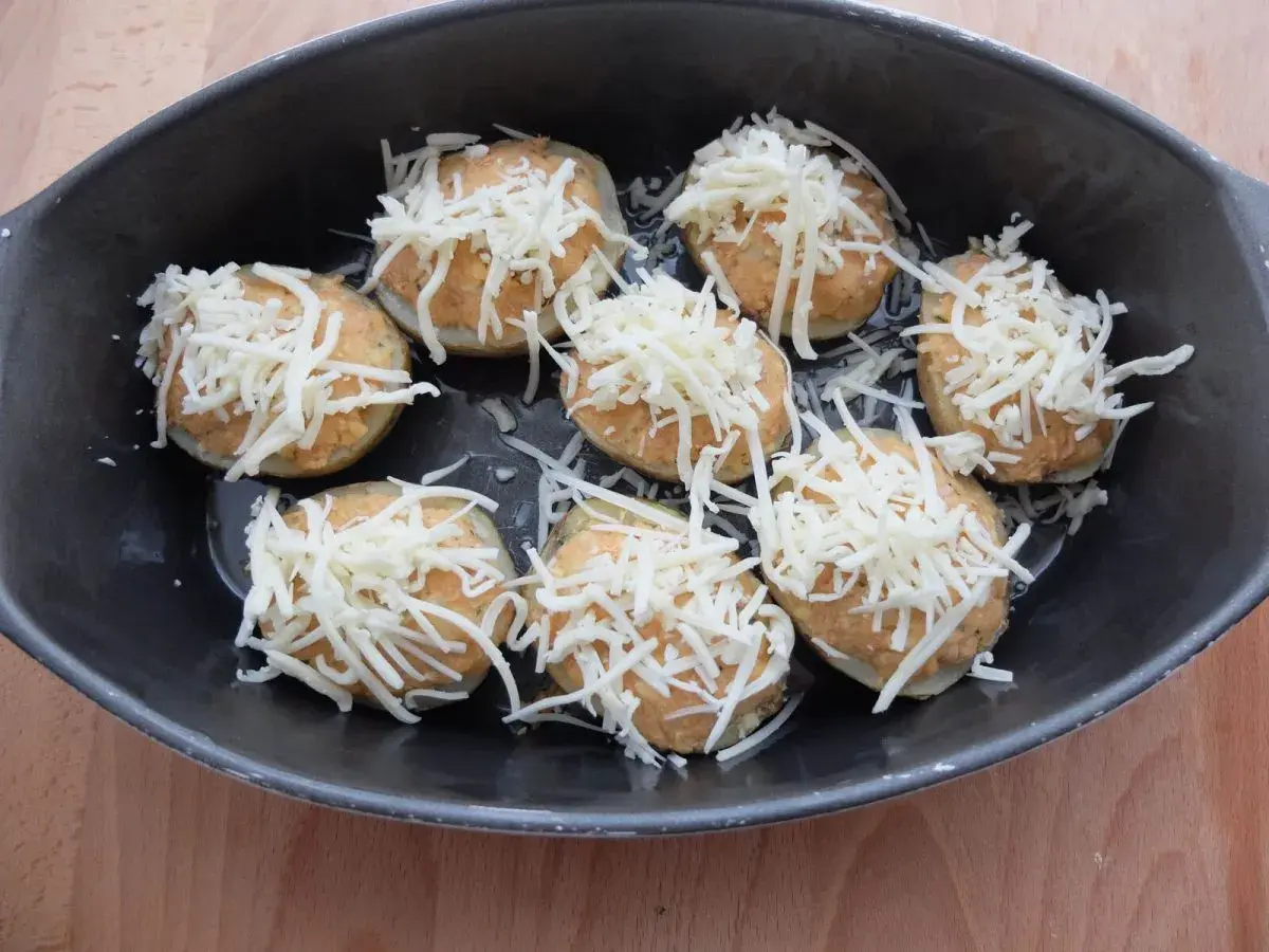Patatas rellenas de atún listas para hornear, cubiertas con queso rallado en una bandeja oscura.