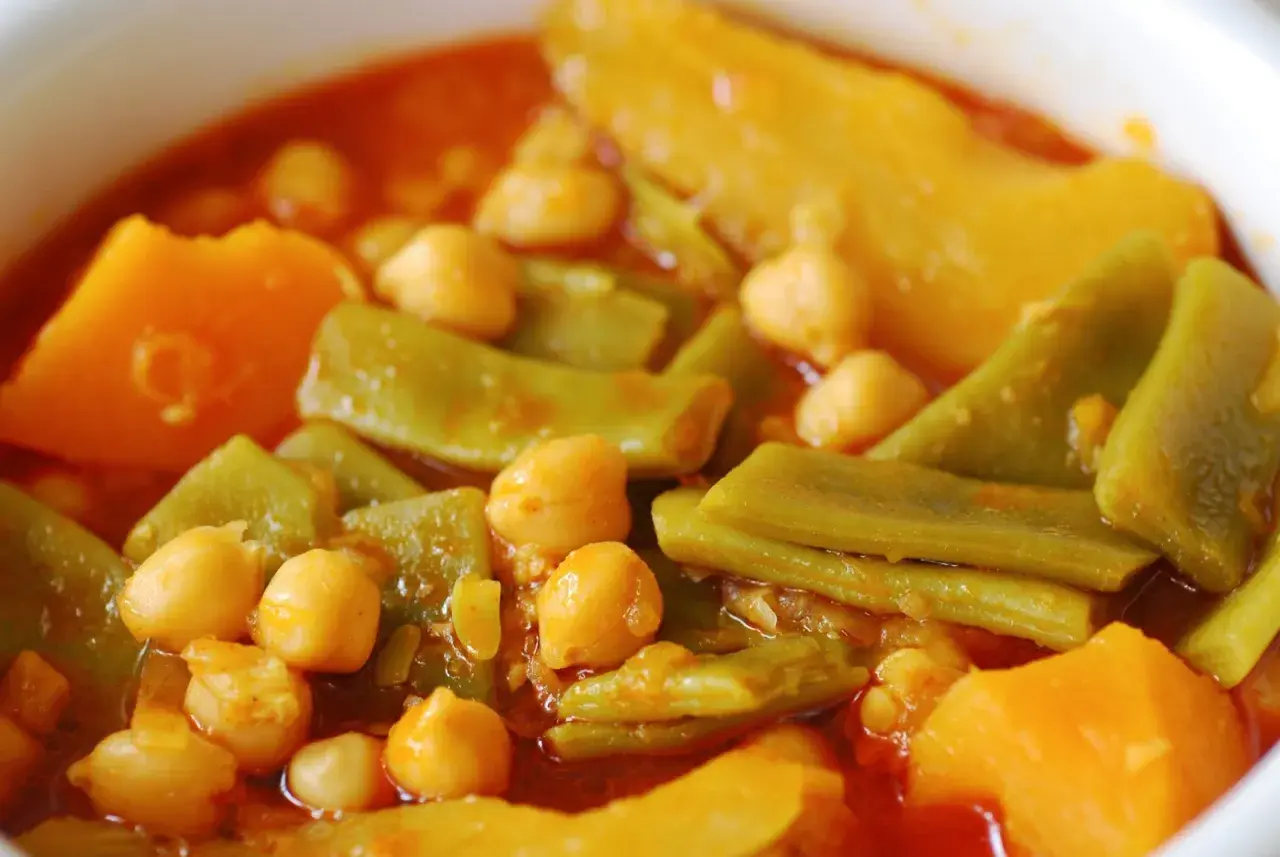 Un plato humeante de olla gitana, con garbanzos, judías verdes y trozos de calabaza en una salsa rojiza.