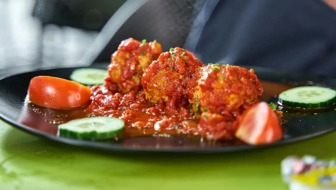 Albondigas con tomate, jugosas y cubiertas de salsa roja, acompañadas de rodajas de pepino y tomate fresco en un plato negro.