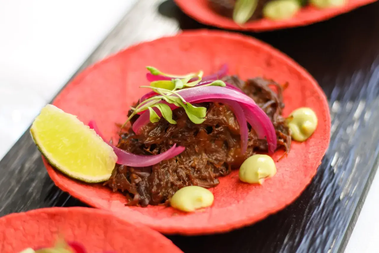 Deliciosos tacos de rabo de toro sobre tortilla roja, con cebolla morada, brotes verdes y un toque de aguacate.