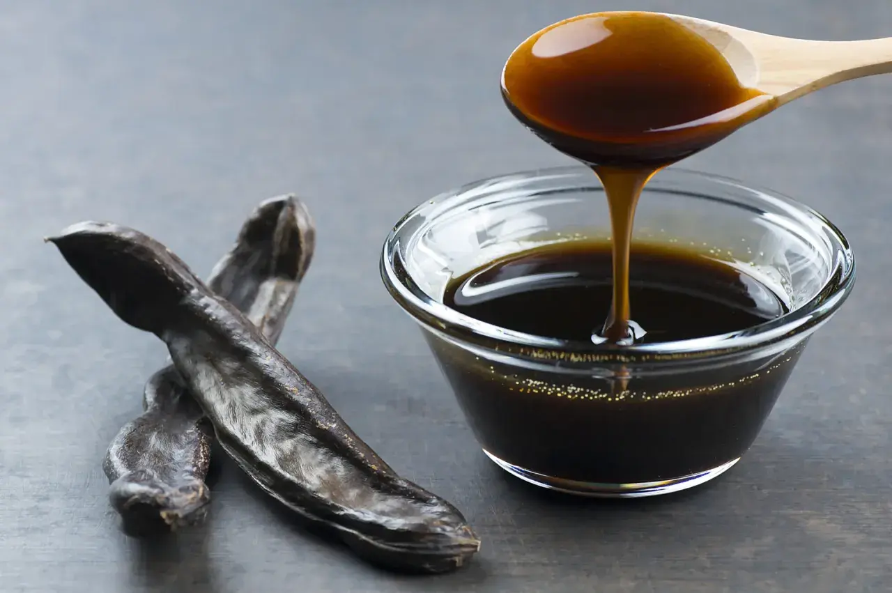 Miel de caña oscura y espesa cayendo de una cuchara de madera a un cuenco de cristal, junto a vainas de algarroba. Descubre sus propiedades.