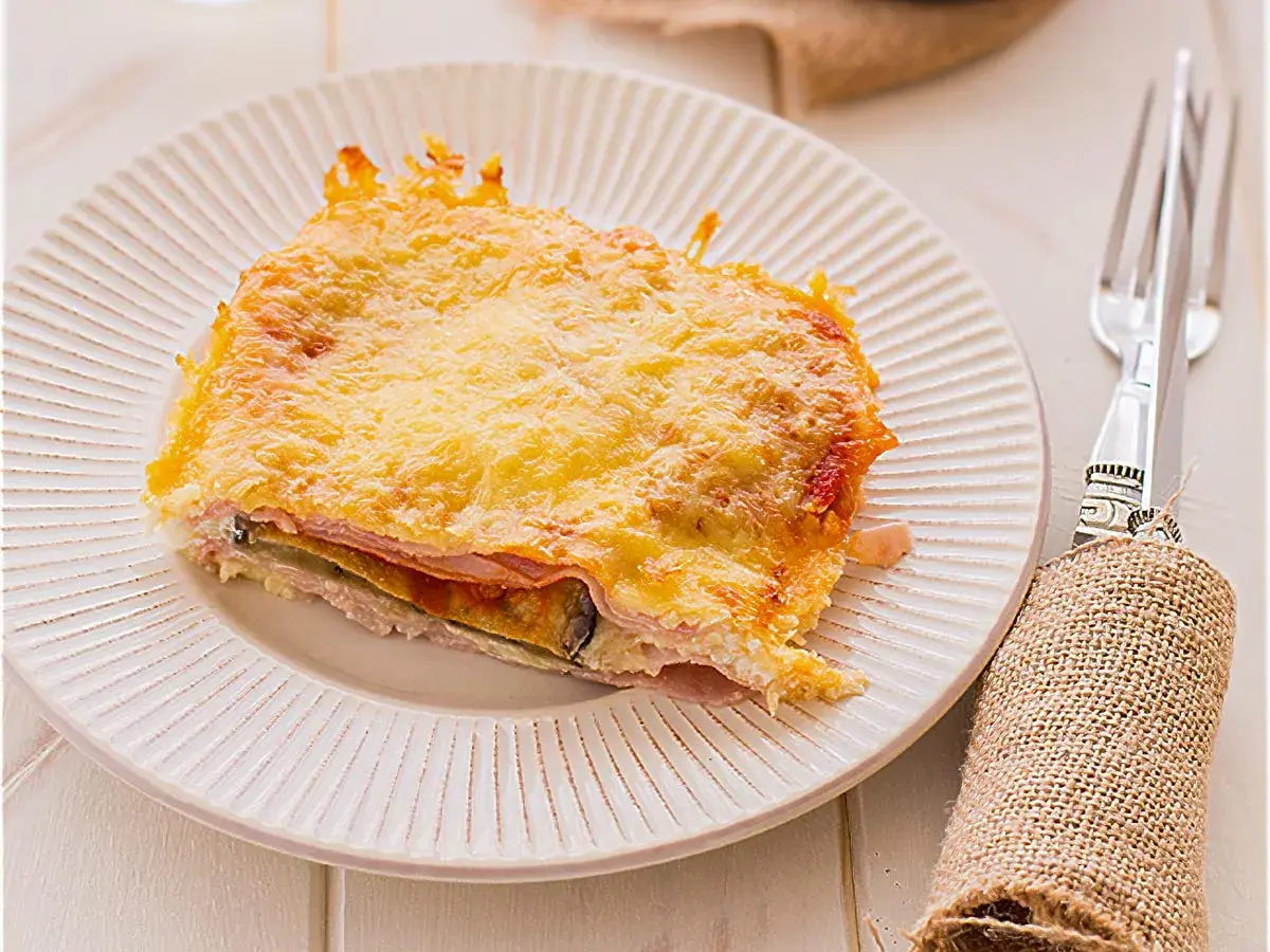 Porción de pastel de berenjenas gratinado con queso, capas de jamón y berenjena, servido en plato blanco.