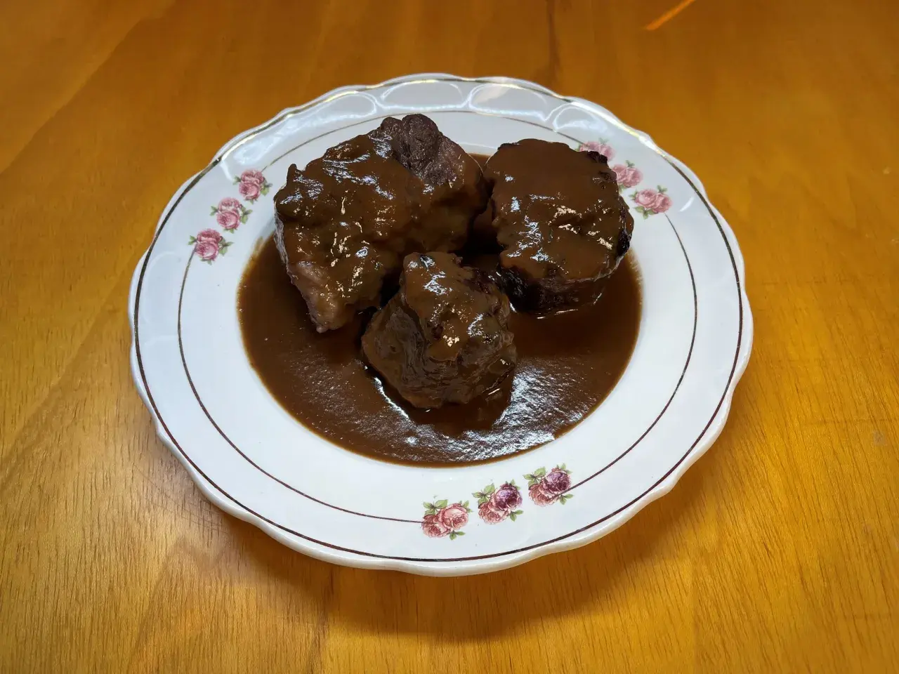 Tres trozos de rabo de toro estofado en una salsa oscura y rica, servidos en un plato blanco con borde dorado y decorado con rosas.