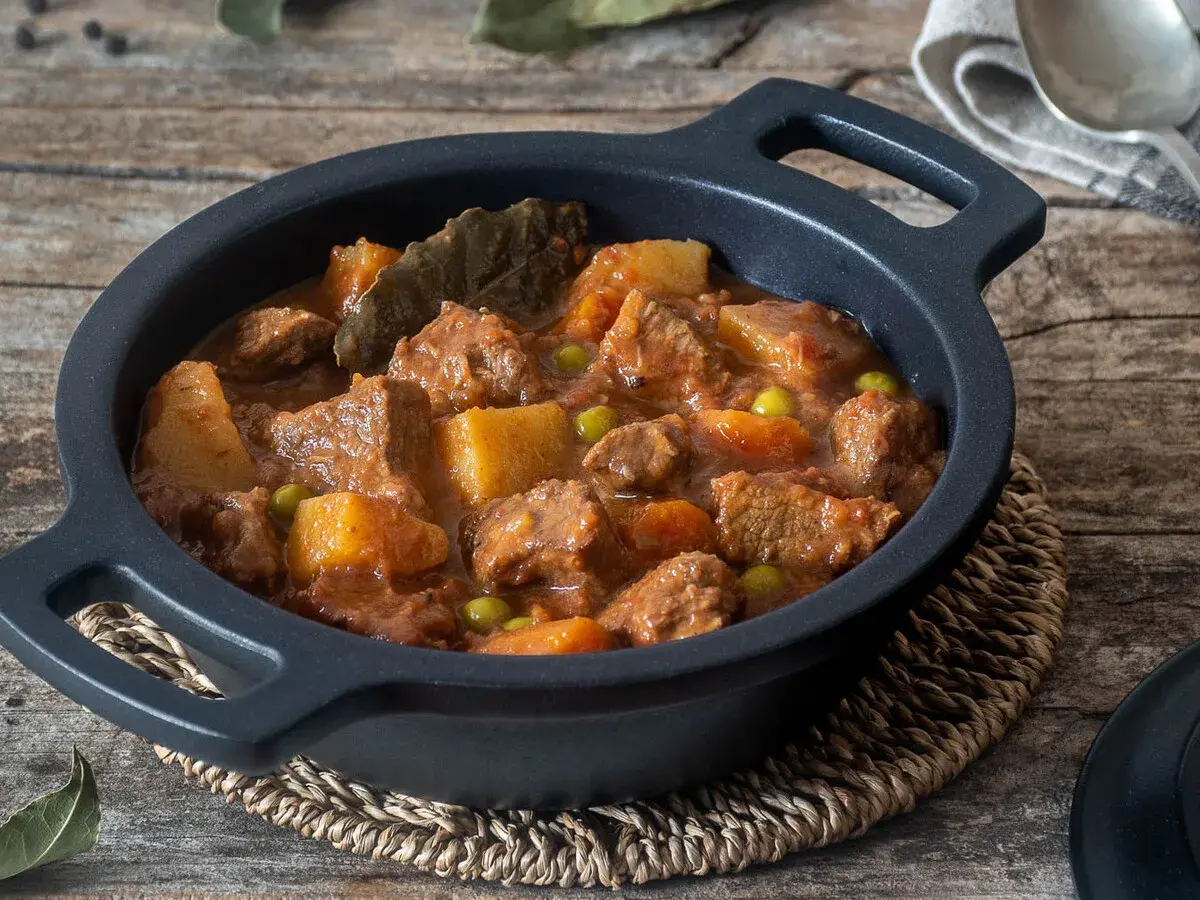 Un reconfortante estofado de ternera con patatas, guisantes y laurel en una cazuela negra sobre madera rústica.