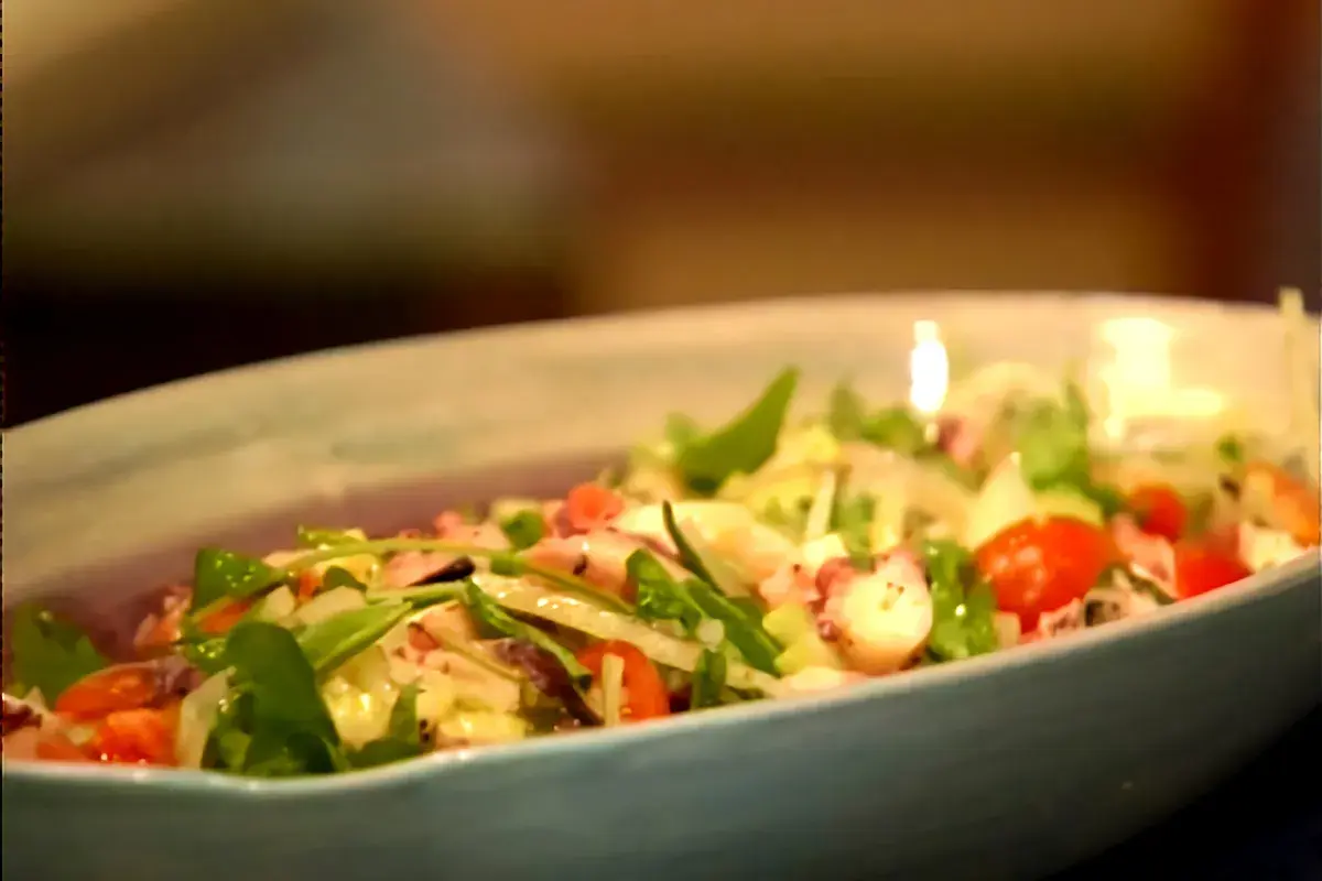 Deliciosa insalata di mare con tentáculos de pulpo, tomates cherry y hojas verdes frescas en un plato azul claro.