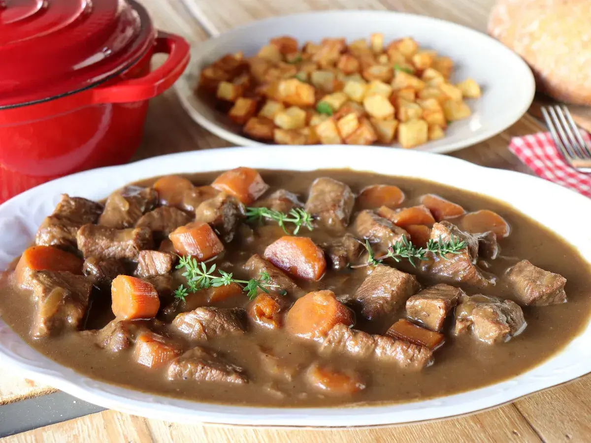 Ternera estofada en salsa con zanahorias y patatas fritas al fondo.
