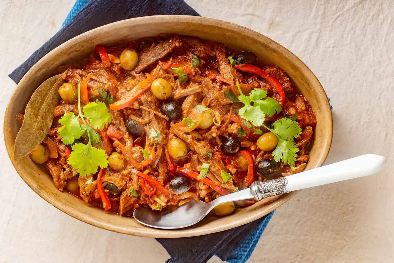 Delicioso plato de ropa vieja con aceitunas verdes y negras, pimientos rojos y cilantro fresco, servido en una cazuela de barro.