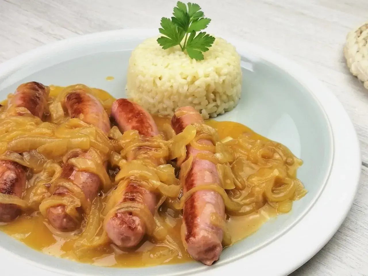 Salchichas al vino con cebolla caramelizada y una porción de arroz blanco, decorado con perejil.