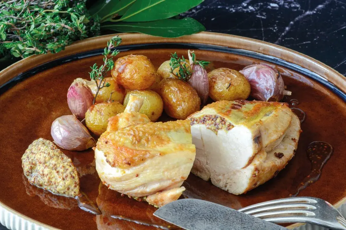 Pollo asado con patatas y ajos, acompañado de una generosa porción de mostaza de grano entero, mostrando varios tipos de mostaza.