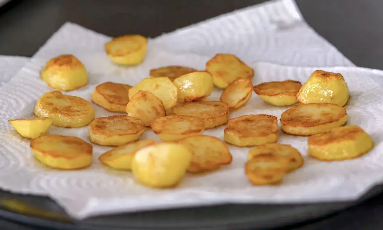 Rodajas de patatas asadas doradas y crujientes, reci&eacute;n salidas de la sart&eacute;n, reposando sobre papel absorbente.
