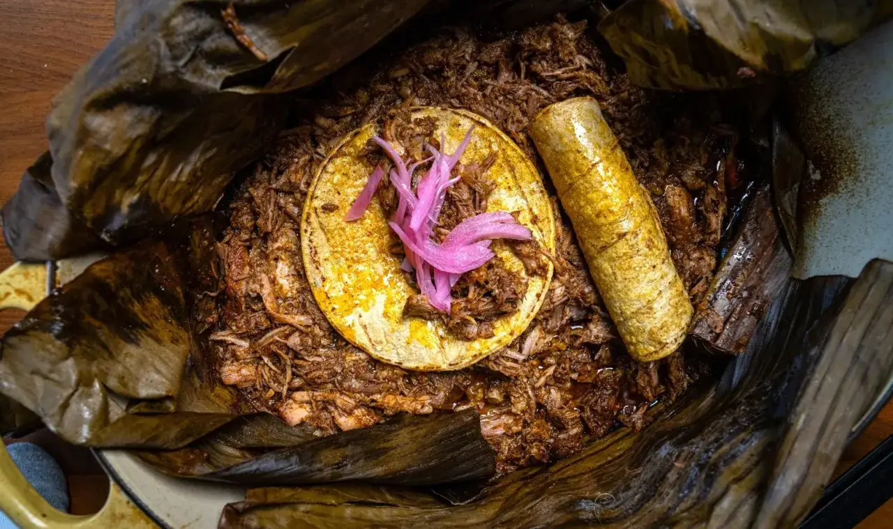 Cochinita pibil que es: cerdo deshebrado en hoja de plátano, servido con tortilla y cebolla morada encurtida.