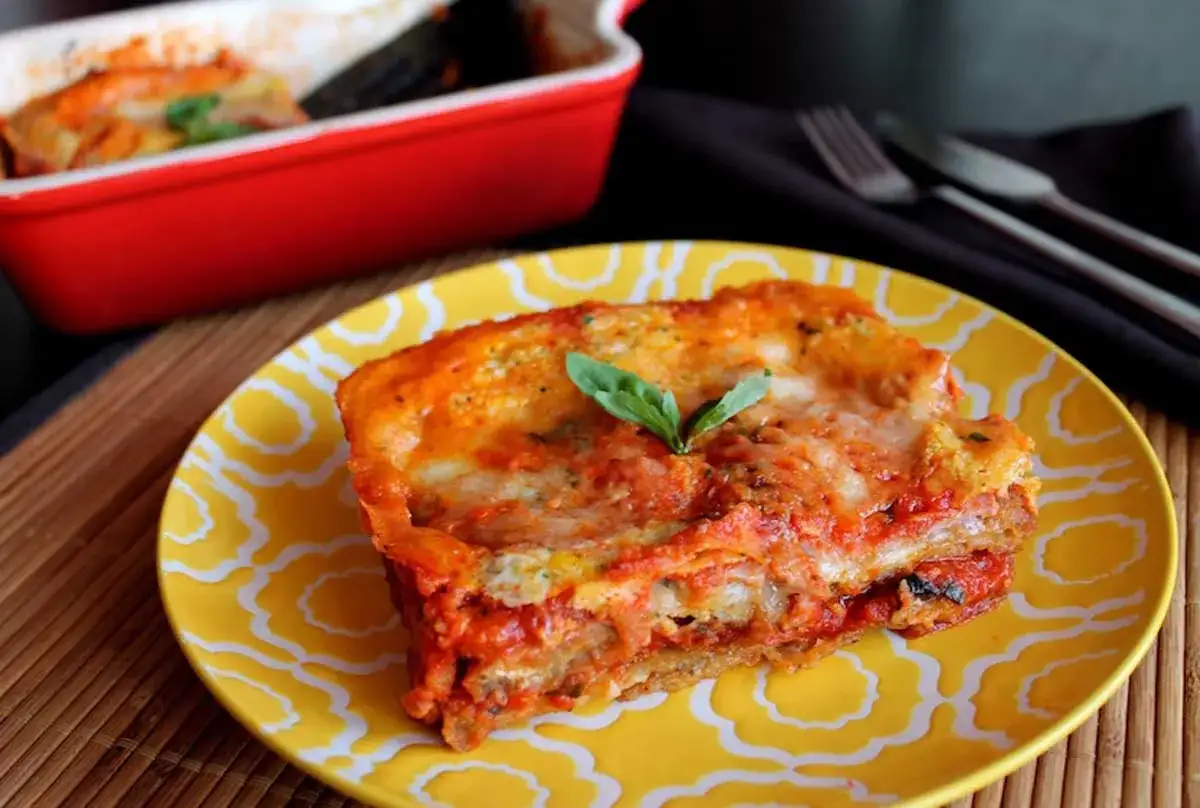 Porci&oacute;n de pastel de berenjenas gratinado con queso y salsa de tomate, decorado con una hoja de albahaca.