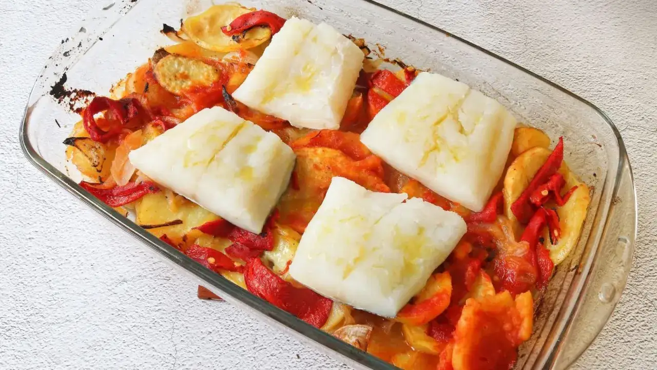 Cuatro porciones de bacalao al horno, jugosas y tiernas, reposan sobre una cama de patatas y pimientos asados en una fuente de cristal.