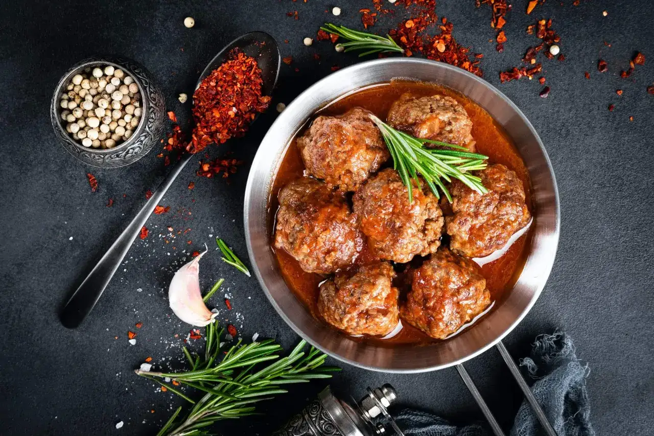 Sabrosas albóndigas con tomate, servidas en un plato metálico, acompañadas de romero, ajo y especias.
