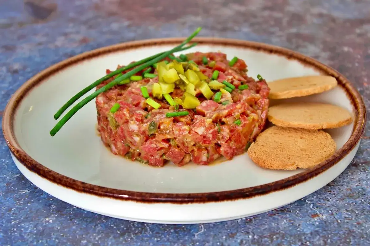 Tartar de salchich&oacute;n picado finamente, adornado con pepinillos y cebollino, servido con tostadas.