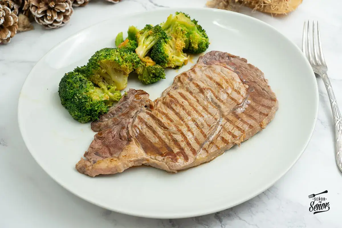 Un jugoso entrecot a la plancha con marcas de parrilla, acompañado de brócoli al vapor en un plato blanco.