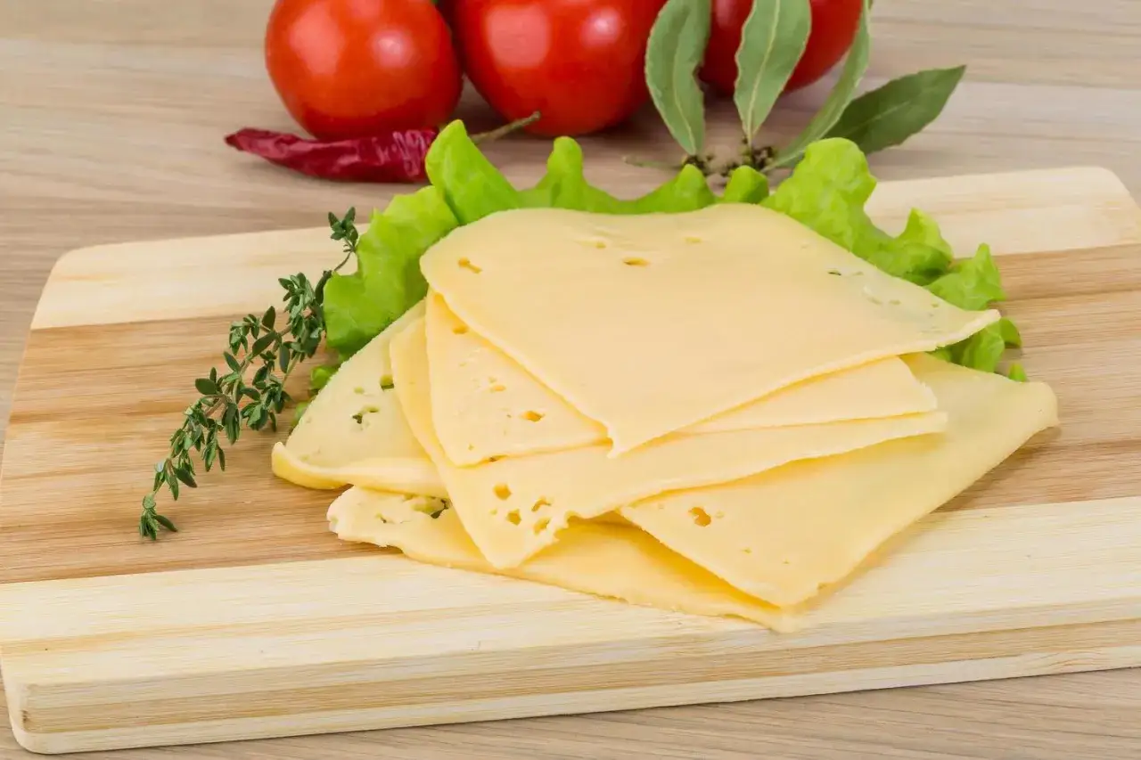 Lonchas de queso cheddar sin lactosa sobre lechuga, con tomates y hierbas al fondo.