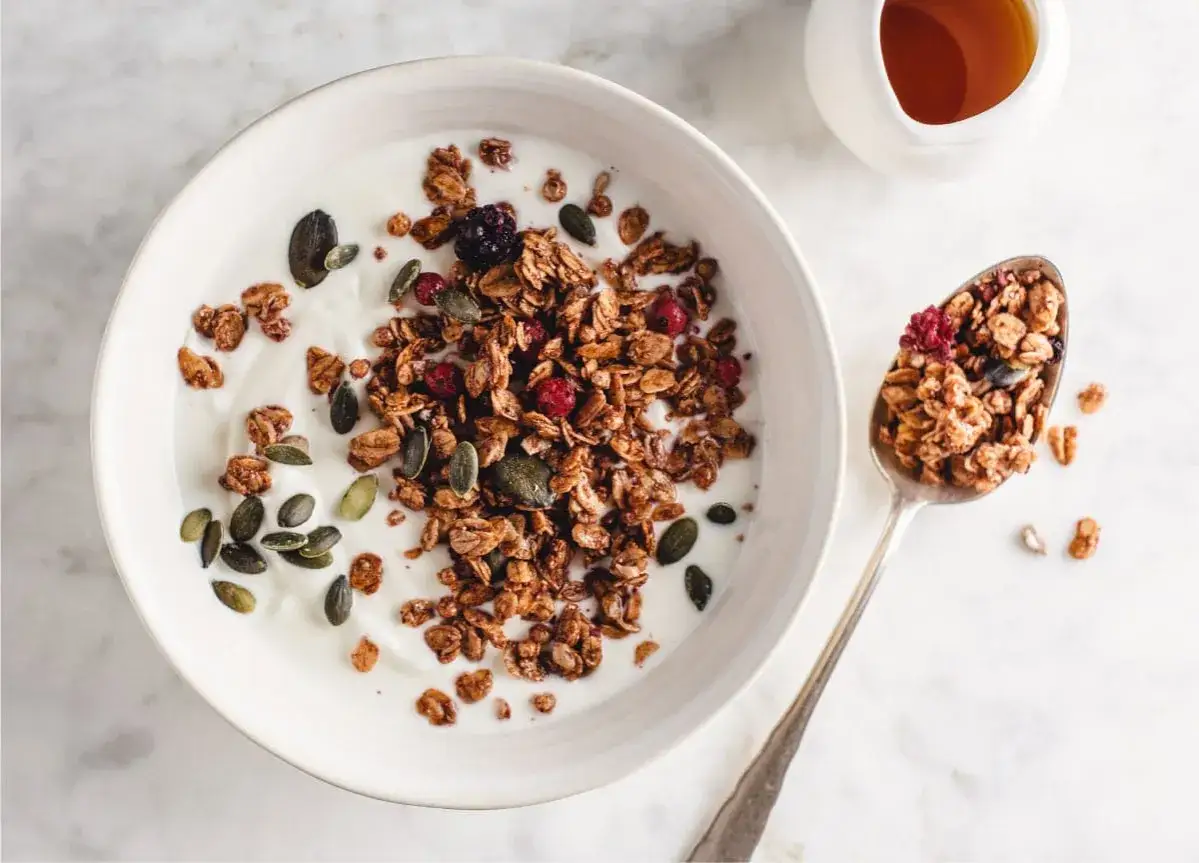 Un tazón de yogur con muesli crujiente, semillas de calabaza y frutos rojos. Una cuchara llena de muesli, ideal para un desayuno saludable.
