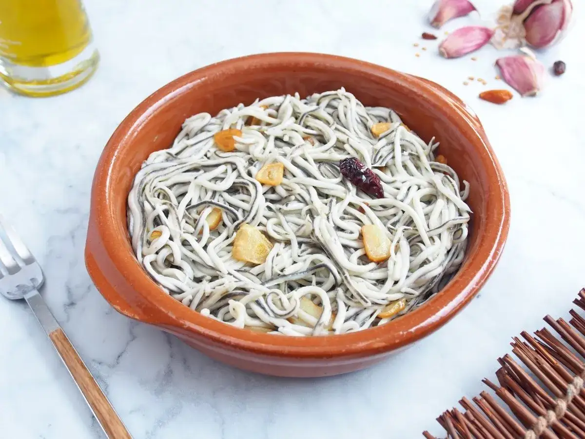 Taz&oacute;n de barro con gulas al ajillo, salteadas con ajo y guindilla. Se ve una botella de aceite de oliva y ajos en el fondo.