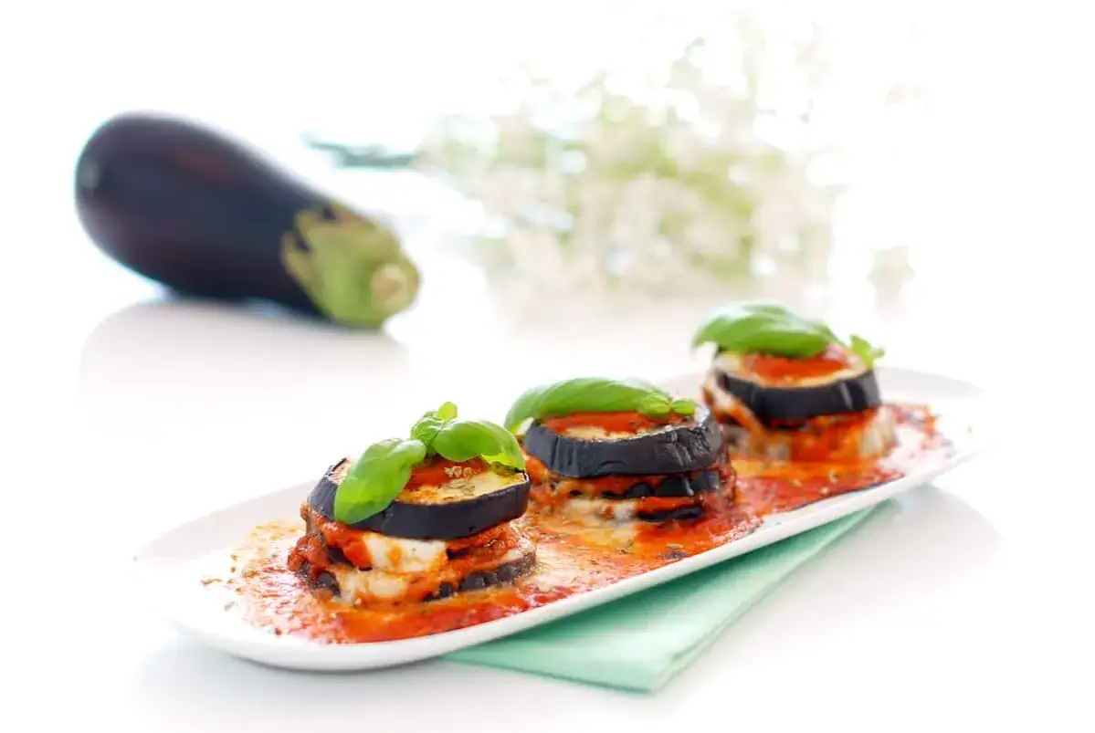 Tres porciones de parmigiana de berenjena, con capas de berenjena, queso y salsa de tomate, decoradas con albahaca fresca.