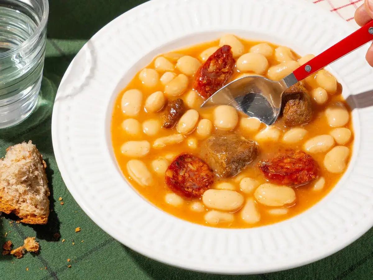 Un plato humeante de judiones de la granja con chorizo y trozos de carne. Una cuchara plateada con mango rojo se hunde en el guiso.